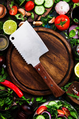 Healthy vegan ingredients layout. Flat-lay of green herbs, tomatoes, cucumbers, spices and oil on brown kitchen table with wooden board and hatchet knife. Top view, copy space.