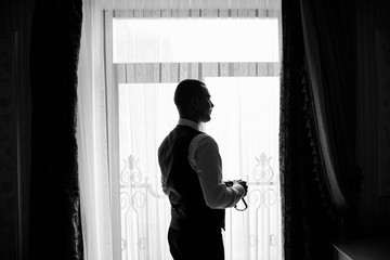Stylish groom near window. Preparation of groom before wedding at home. Sexy man posing. Groom getting ready in the room for wed day.