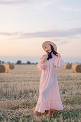 Attractive girl in dress and strw hat is walkinkg on field at dawn