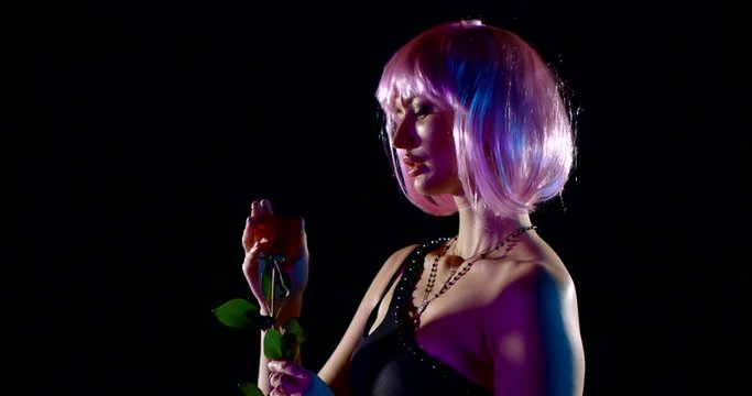portrait of a woman with pink hair in black underwear in the dark. she holds a red rose, tears off the petals and sprinkles herself. contoured blue and purple light