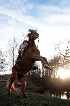 Pferd steigt im Sonnenuntergang vor einem See währen die Reiterin ein Schwert in der Hand hält