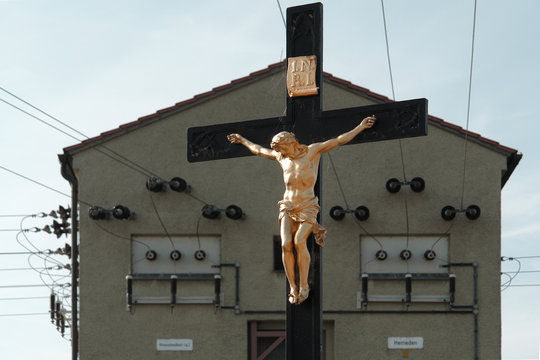A crucifix  - golden sculpture of Jesus Christ hanging on a wooden cross - in front of a transformer station in Herrieden, Bavaria