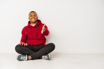 Young latin man sitting on the floor isolated stretching hand at camera in greeting gesture.