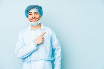 Young arabian surgeon man isolated against on a blue background smiling and pointing aside, showing something at blank space.
