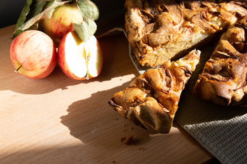 The homemade apple pie and fresh apples on a wooden table in the kitchen, sliced piece of pie