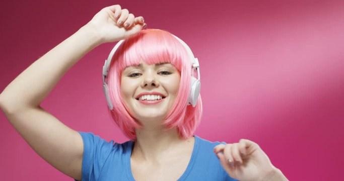 Close up of attractive cheerful young smiled woman in pink short wig and headphones dancing joyfully. Portrait of beautiful hapy gir listening to music and laughing on bright background wall.