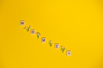 Top view word women spelled in wooden blocks on yellow background with flowers between them. Love, 8 march background. Gift, greeting, compliment concept. Copy space. place for text.