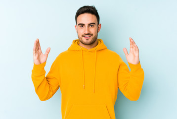 Young caucasian man isolated on blue background holding something little with forefingers, smiling and confident.