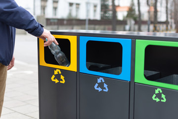 Hand of man throwing plactic bottle into trash bin for plastic waste. Modern colorful refuse bins for separate trash. - obrazy, fototapety, plakaty