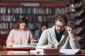 Mature bearded man in casual clothing and eyeglasses sitting at table with lots of book at public library. Serious male searching for information. Education concept.