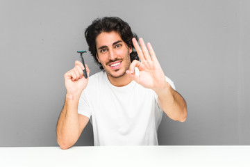 Young man holding a razor blade cheerful and confident showing ok gesture.
