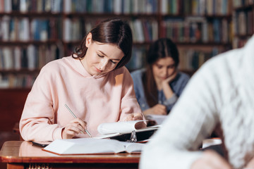 Charming brunette student in pink hoodie studying at university library and taking notes. Young lady preparing for exams among heaps of open books