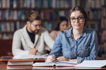 Young female student with hair knot in eyeglasses looking at camera while sitting at table with open books. Pretty woman in jeans shirt studying at university library