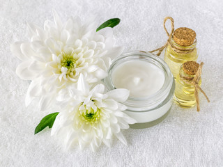 Fototapeta na wymiar Face cream in an open glass jar, two bottles of chrysanthemum essential oil and two white chrysanthemums on a white terry towel. Beauty, skincare and cosmetology.