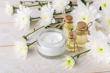 Obraz na płótnie Canvas Face cream in an open glass jar, three bottles of chrysanthemum essential oil and white chrysanthemum flowers on a wooden table. Beauty, skincare and cosmetology.