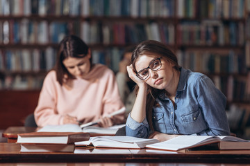 Beautiful tired student in jeans shirt and eyeglasses sitting at table with open books and looking at camera. Pretty lady with brown hair gaining knowledge at university library