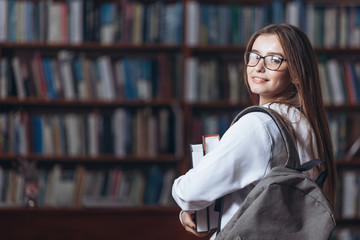 Cheerful female student with long brown hair standing in university library with books in hands. Young woman in casual clothing and eyeglasses smiling and looking at camera.