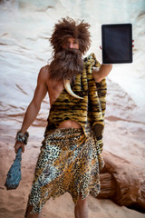 Modern caveman holding the blank screen of a tablet outdoors in a weathered rock cave; selective focus on the tablet in the foreground