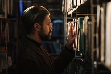 Side view of mature bearded man in formal clothing standing among shelves at public library and choosing book to read. Confident male spending time for self education