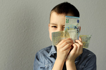 kid holds euro banknotes in his hands near his face, looks with one eye, concept of pocket money,...
