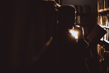 Side view of bearded reader leaning on bookshelf and holding open book at library with dark lights....