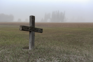 As simple wooden cross marking a lonely grave remote from the rest of the headstones.  The setting is a foggy morning creating a gloomy setting.