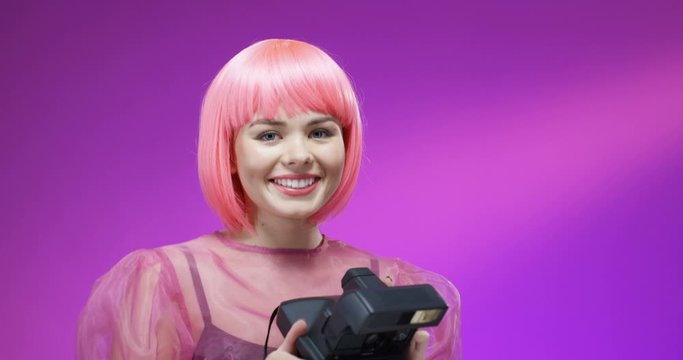 Close up of Caucasian young beautiful and stylish woman in pink wig taking photos with photocamera polaroid and smiling. Pretty girl taking pictures on instant photo camera on bright background.