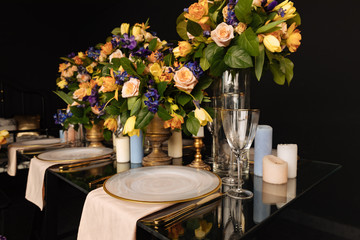 a large flower arrangement with candles on a transparent glass table with white gilded plates and gilded forks, knives and glasses. floral table decoration with expensive dishes