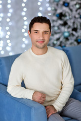Handsome man wearing white sweater sitting on the lue sofa. Comfort and relaxation on gray...