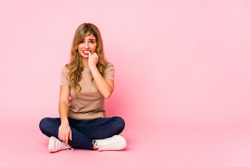 Young blonde caucasian woman sitting on a pink studio biting fingernails, nervous and very anxious.