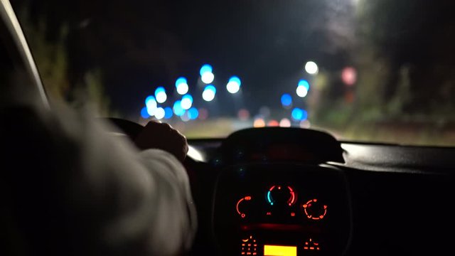 Illuminated car dashboard, automobile interior. Driver turning steering wheel, accelerating speed and driving along illuminated with neon lights lamps steets at night, glare in the window. Driver