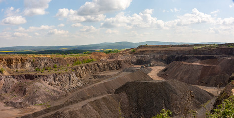 View of basalt (brown rock) quarry with rock exploitation during summer - blue sky with clouds, open-pit plant and village and background