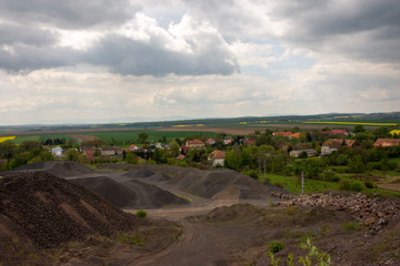 Fototapeta na wymiar View of basalt (brown rock) quarry with rock exploitation during summer - blue sky with clouds, open-pit plant and village and background