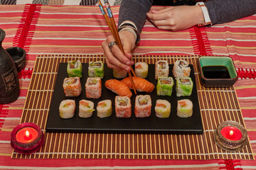 Happy lady holds a sushi roll on the chopsticks, looks at delicious Japanese food and smiles, over a table sit on a sofa