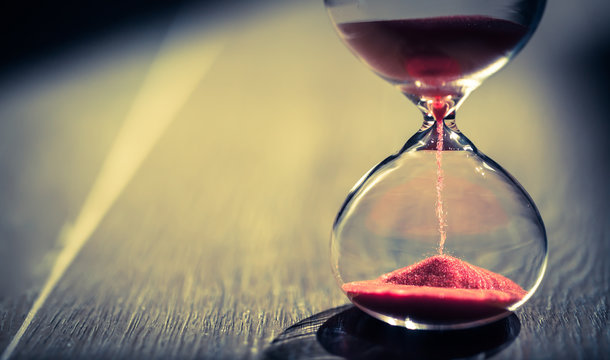 Red sand running through the bulbs of an hourglass measuring the passing time in a countdown to a deadline, on a wooden floor background with copy space. Under sunlight. 
