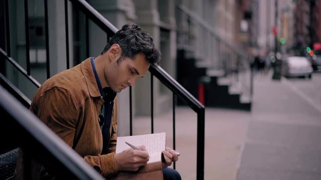Latin male in trendy outfit writing information in personal planner while sitting at urban setting in city environment, clever man making notes in knowledge textbook during free time outdoors