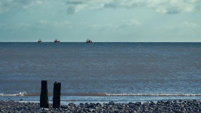 Three distant fishing boats working in the Irish Sea in Leinster, with gentle waves lapping on a pebble beach.