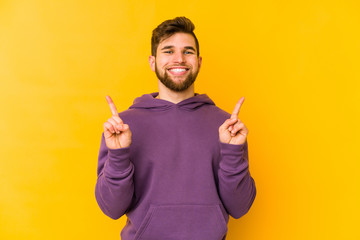 Young caucasian man isolated on yellow background indicates with both fore fingers up showing a blank space.