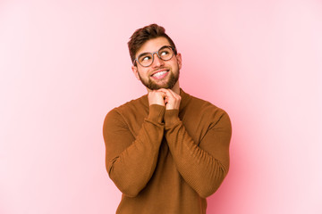 Young caucasian man isolated on pink background keeps hands under chin, is looking happily aside.