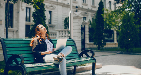 Cheerful red haired caucasian girl looking up in the sky while sitting on the bench and working at the computer