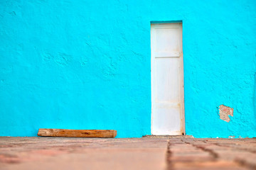 Narrow white door in a simple blue wall. Minimal Mediterranean village architecture detail.