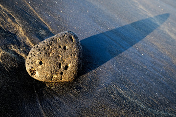 Large black basalt lava boulder at sunset on a black volcanic sand beach.