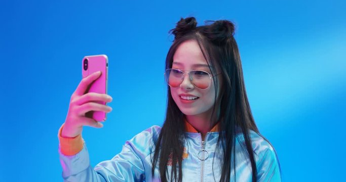 Chinese young pretty woman in stylish look posing to smartphone camera. Beautiful Asian girl taking selfie photo with phone on blue bright background. Female making selfies photos.
