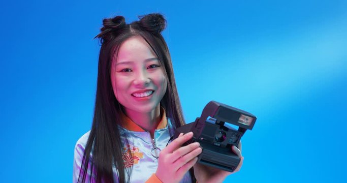 Close up of Asian young beautiful and stylish woman taking photos with photocamera polaroid and smiling. Pretty Chinese girl taking pictures on instant photo camera on bright blue background.