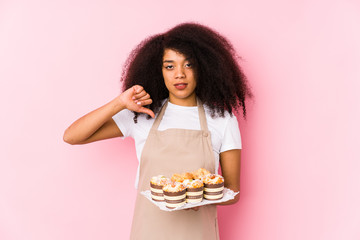 Young afro pastry maker woman holding a cupcakes isolatedYoung afro baker woman showing a dislike gesture, thumbs down. Disagreement concept.