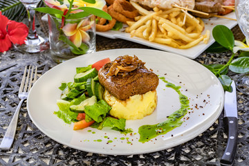 Grilled steak, caramelized onions, coffee sauce, and green mint sauce, mashed potatoes and vegetable salad on a white plate, , blurred restaurant background