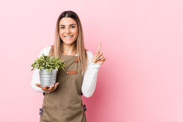 Young gardener woman holding a plant smiling and pointing aside, showing something at blank space.