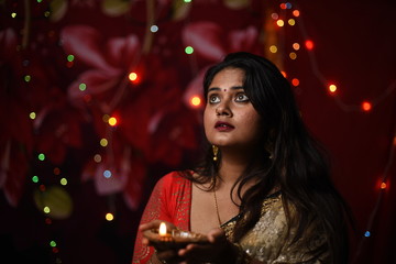 An young and beautiful Indian Bengali woman in Indian traditional dress is holding a Diwali diya/lamp in her hand standing in front of colorful bokeh lights. Indian lifestyle and Diwali celebration