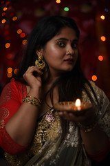 An young and beautiful Indian Bengali woman in Indian traditional dress is holding a Diwali diya/lamp in her hand standing in front of colorful bokeh lights. Indian lifestyle and Diwali celebration