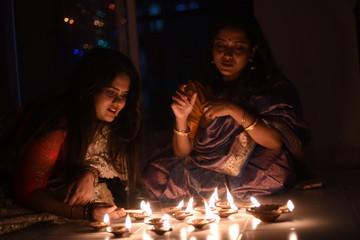 Two beautiful Indian Bengali women in Indian traditional dress are lighting Diwali diya/lamps sitting on the floor indoor in darkness on Diwali evening. Indian lifestyle and Diwali celebration